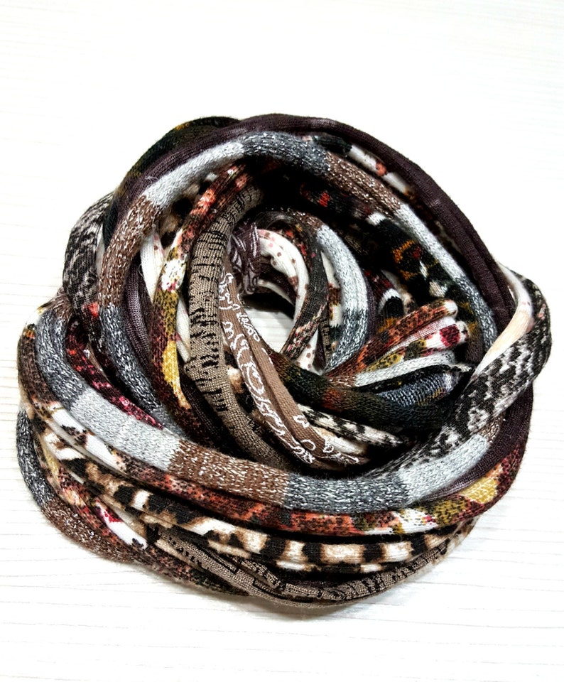 May include: A circular infinity scarf featuring a multi-patterned knit fabric in shades of brown, black, white, and gray. The textile displays various textures and prints, including animal-inspired motifs and geometric patterns, coiled into a dense, layered loop.