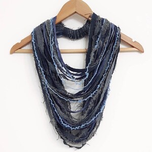 May include: A multi-strand infinity scarf constructed from various strips of dark blue, charcoal gray, and light blue denim fabric. The strips feature frayed, raw edges and are layered to create a textured, cascading effect around the neck.