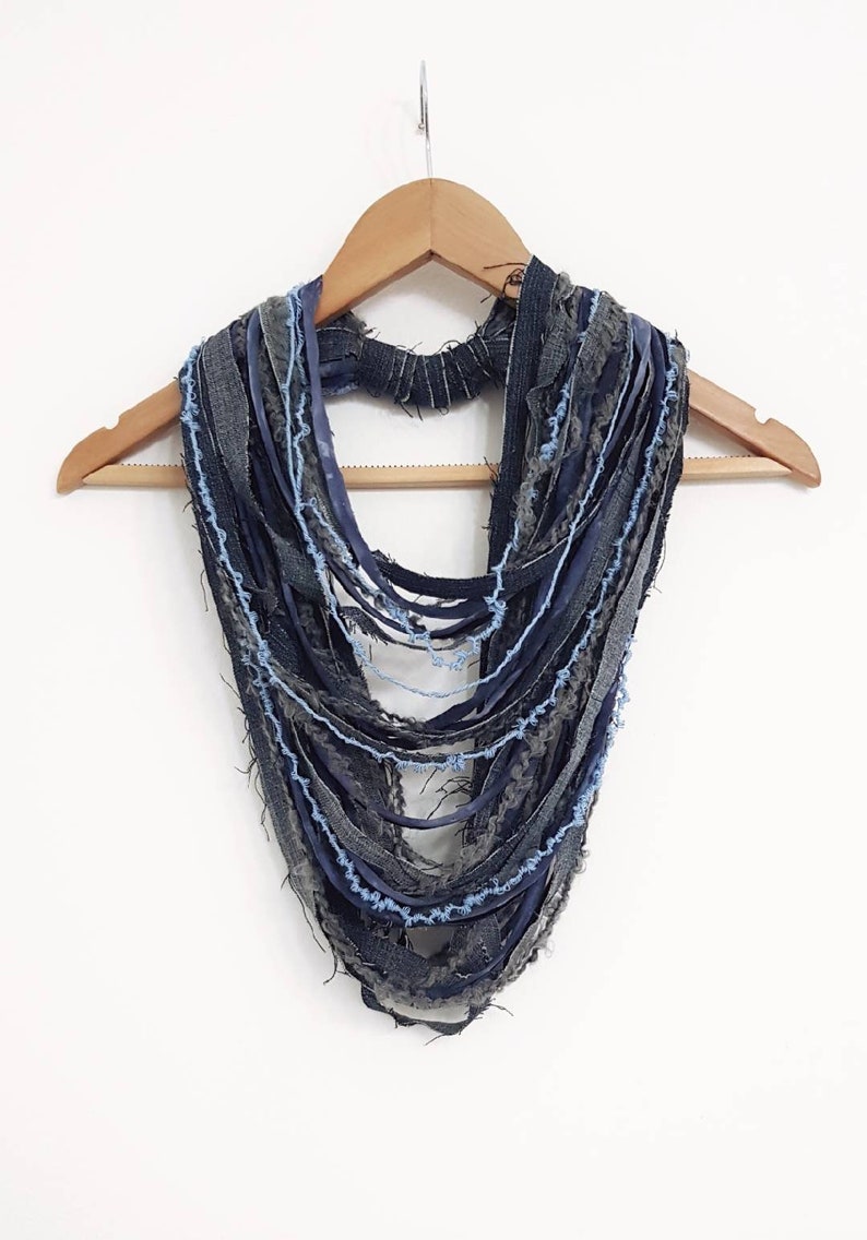 May include: A multi-strand infinity scarf constructed from various strips of dark blue, charcoal gray, and light blue denim fabric. The strips feature frayed, raw edges and are layered to create a textured, cascading effect around the neck.