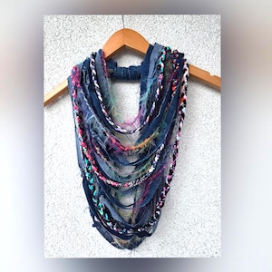 Puede incluir: Bufanda infinita de mezclilla azul oscuro hecha a mano con detalles coloridos. La bufanda presenta tiras de mezclilla y hilo trenzado en tonos rosa, morado, naranja y blanco. Cuelga de una percha de madera sobre un fondo blanco texturizado.