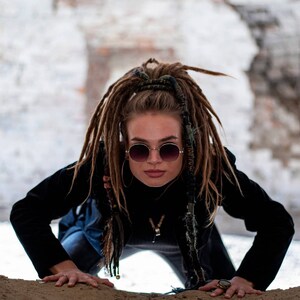 K&ouml;nnte beinhalten: Eine Person mit langen braunen Dreadlocks in einem hohen Pferdeschwanz, tr&auml;gt runde, verlaufende Sonnenbrillen mit d&uuml;nnem Goldrahmen, ein schwarzes Langarmshirt und eine schwarze Jacke. Die Person hat ein kleines silbernes Gesichtspiercing und Ringe.