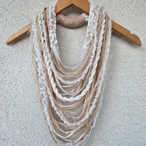 Op de afbeelding: Een handgemaakte sjaal in bohemien-stijl in cr&egrave;me, beige en bruintinten, gepresenteerd op een houten hanger. De sjaal heeft een mix van texturen, waaronder gevlochten elementen, kanten randen en vloeiende linten.