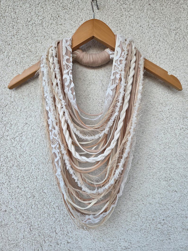 Op de afbeelding: Een handgemaakte sjaal in bohemien-stijl in cr&egrave;me, beige en bruintinten, gepresenteerd op een houten hanger. De sjaal heeft een mix van texturen, waaronder gevlochten elementen, kanten randen en vloeiende linten.