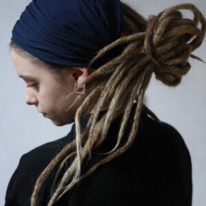K&ouml;nnte beinhalten: Eine Person tr&auml;gt ein marineblaues Kopftuch und ein schwarzes Oberteil. Die Person hat lange, hellbraune Dreadlocks. Die Person tr&auml;gt auch quadratische Ohrringe. Der Hintergrund ist ein schlichtes Wei&szlig;.