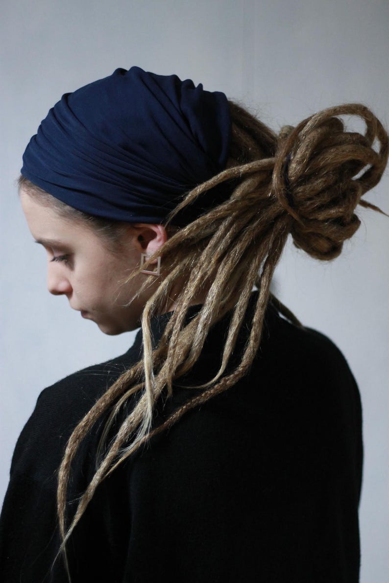 K&ouml;nnte beinhalten: Eine Person tr&auml;gt ein marineblaues Kopftuch und ein schwarzes Oberteil. Die Person hat lange, hellbraune Dreadlocks. Die Person tr&auml;gt auch quadratische Ohrringe. Der Hintergrund ist ein schlichtes Wei&szlig;.