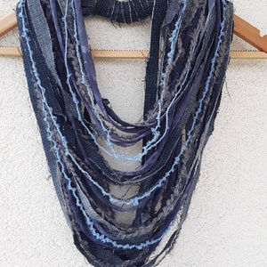May include: A multi-strand infinity scarf constructed from various strips of dark blue denim, grey fabric, and light blue yarn. The design features frayed edges and a layered, cascading appearance, with a central band holding the strands together.