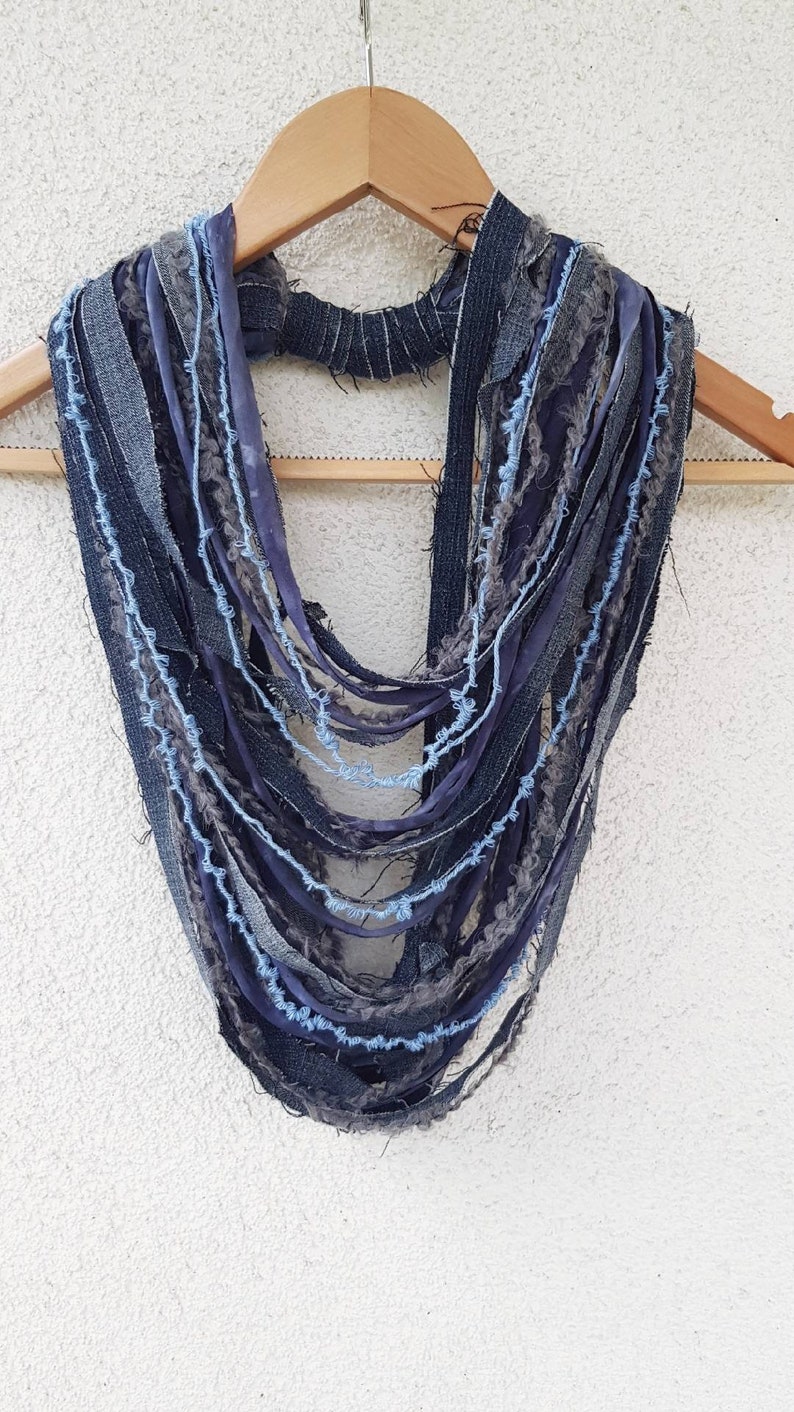 May include: A multi-strand infinity scarf constructed from various strips of dark blue denim, grey fabric, and light blue yarn. The design features frayed edges and a layered, cascading appearance, with a central band holding the strands together.