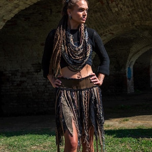 May include: A woman wearing a black crop top, a brown and black skirt, and a layered necklace. The skirt has a fringed design. The background features a brick archway and green grass.