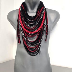May include: A layered necklace displayed on a mannequin. The necklace features strands of black, red, and patterned fabric. Braided red strands and tassels add detail. The overall design is bold and modern.