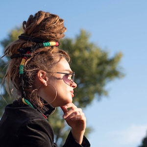 May include: A woman with long brown dreadlocks wearing sunglasses and a black jacket. She is wearing a colorful beaded headband and has a large hoop earring in her left ear.