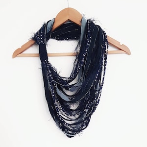 Peut inclure: Une écharpe infinie bleu foncé composée de multiples bandes de tissu effiloché. L'écharpe présente un mélange de textures bleu marine uni et bleu clair façon denim, avec certaines bandes arborant un petit motif à pois blancs sur l'ensemble.