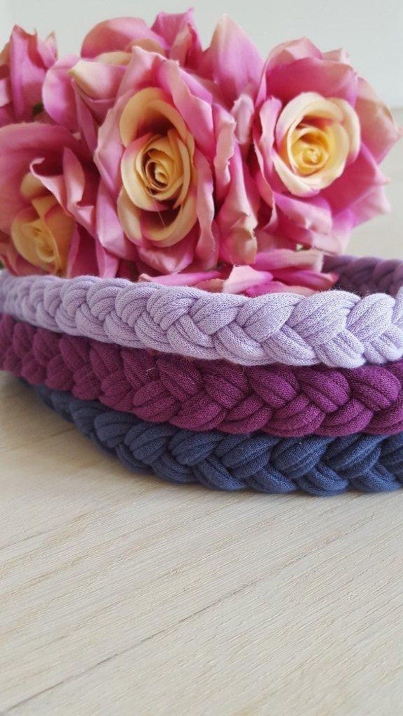 May include: Three braided headbands in light purple, deep purple, and dark blue fabric, stacked horizontally. A cluster of pink and cream-colored roses rests directly behind the headbands, adding a floral element to the composition.