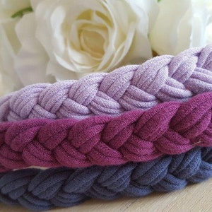 May include: Three braided headbands stacked together, featuring a light lavender, a deep magenta, and a dark slate blue color. Each headband is constructed from a soft, fabric-like material with a consistent, repeating woven braid pattern.