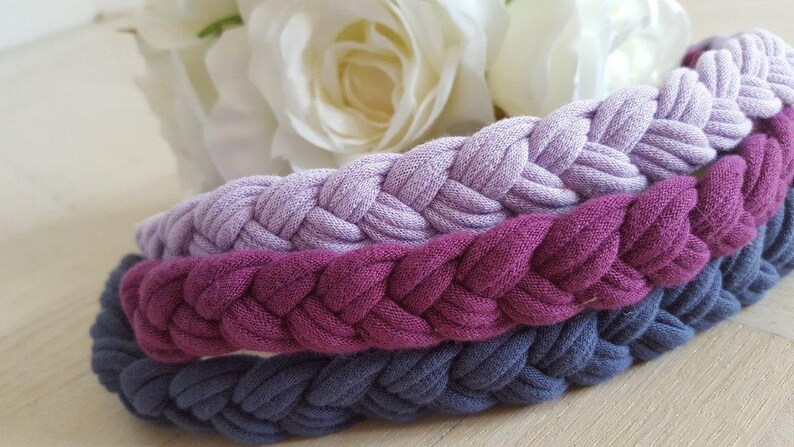 May include: Three braided headbands stacked together, featuring a light lavender, a deep magenta, and a dark slate blue color. Each headband is constructed from a soft, fabric-like material with a consistent, repeating woven braid pattern.