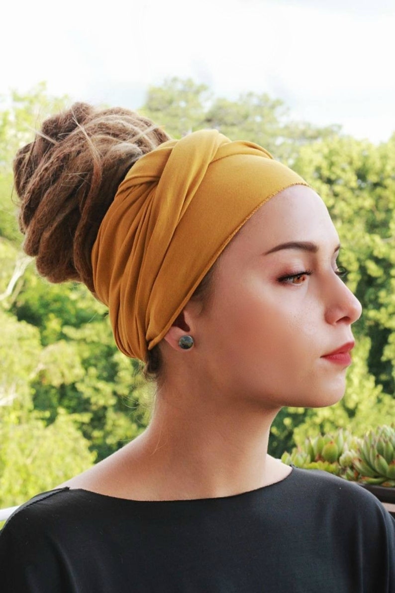 May include: A woman wearing a mustard yellow head wrap. The head wrap is wrapped around the head and tied at the top. The woman is wearing a black top and a pair of small, dark earrings. Her hair is styled in dreadlocks.