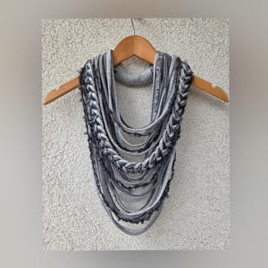 Puede incluir: Bufanda infinita gris con diseño trenzado y detalles de flecos. La bufanda está hecha de tela suave y cuelga de una percha de madera. La bufanda tiene múltiples capas y una abertura circular para el cuello.