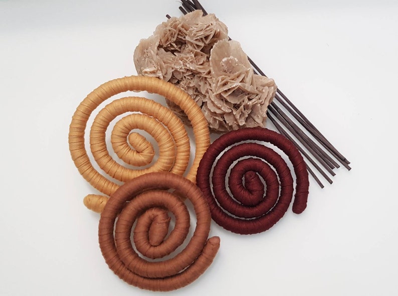 May include: Three coiled, fabric-wrapped circular objects in shades of tan, medium brown, and deep burgundy. Accompanying these are a cluster of light beige, crystalline desert rose mineral formations and a bundle of thin, dark brown wooden sticks.