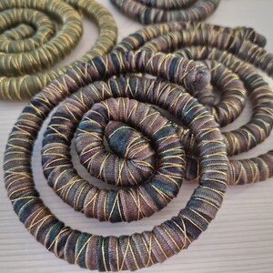 K&ouml;nnte beinhalten: Mehrere kreisf&ouml;rmige Spiralen aus stoffumwickelter Kordel. Die Spiralen weisen ein variierendes Muster in Dunkelbraun, Schwarz und Dunkelgr&uuml;n auf, sind fest gewickelt und mit einem d&uuml;nnen, metallisch goldenen Faden diagonal umwunden.