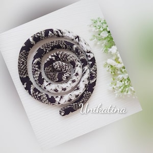 May include: A coiled fabric rope with a snake-like pattern in black, white, and gray. The rope is arranged in a spiral on a white surface, with a sprig of white flowers to the right. The text "Unikatina" is visible.