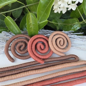 Puede incluir: Una colecci&oacute;n de objetos flexibles tipo cord&oacute;n envueltos en tela, con tres formas de espiral en marr&oacute;n oscuro, terracota y beige, junto a tres piezas largas, rectas y dobladas en colores a juego, dispuestas sobre una superficie blanca.