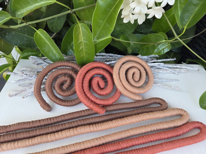 Puede incluir: Una colecci&oacute;n de objetos flexibles tipo cord&oacute;n envueltos en tela, con tres formas de espiral en marr&oacute;n oscuro, terracota y beige, junto a tres piezas largas, rectas y dobladas en colores a juego, dispuestas sobre una superficie blanca.