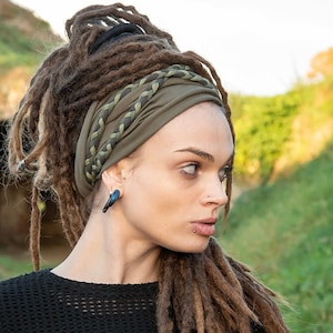 Op de afbeelding: Een olijfgroene stoffen hoofdband met een gevlochten detail aan de voorzijde. De hoofdband wordt gedragen over lange bruine dreadlocks, waarbij de stof is verzameld in een brede, zachte band die over het voorhoofd ligt en het hoofd omsluit.