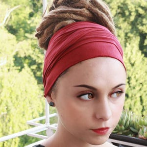 May include: A woman wearing a deep red headband. The headband wraps around the head, covering the forehead and ears. The woman has brown dreadlocks and is wearing a black top. She has a small, dark earring in her left ear.