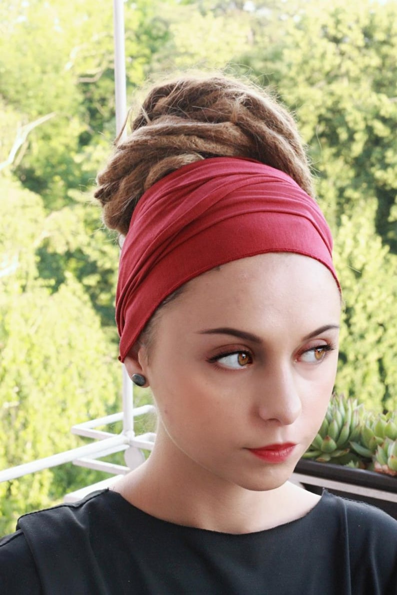 May include: A woman wearing a deep red headband. The headband wraps around the head, covering the forehead and ears. The woman has brown dreadlocks and is wearing a black top. She has a small, dark earring in her left ear.