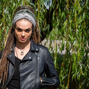 May include: A person wearing a black leather jacket and a turban with shades of gray and green. The person has long dreadlocks and is wearing a necklace with a pendant. The background features green foliage.