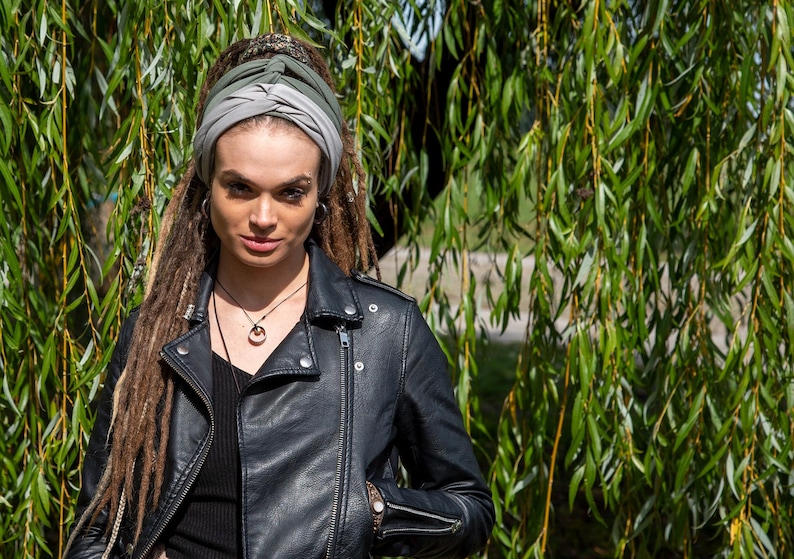May include: A person wearing a black leather jacket and a turban with shades of gray and green. The person has long dreadlocks and is wearing a necklace with a pendant. The background features green foliage.