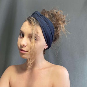 May include: A navy blue headband is worn on the head. The headband is made of a soft fabric and is wrapped around the head. The hair is pulled back and up in a bun. The background is a solid gray color.