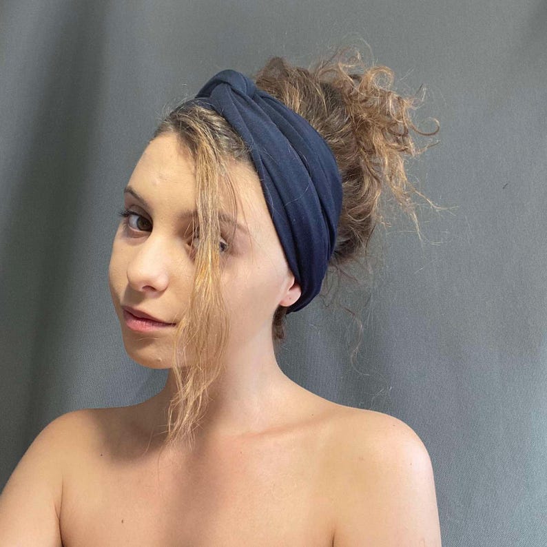 May include: A navy blue headband is worn on the head. The headband is made of a soft fabric and is wrapped around the head. The hair is pulled back and up in a bun. The background is a solid gray color.