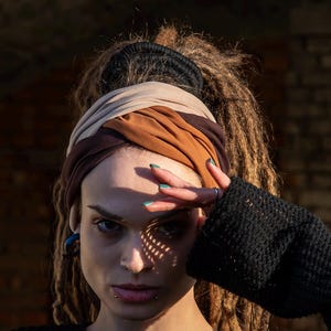 Könnte beinhalten: Eine Frau mit Dreadlocks trägt ein braunes, beiges und schwarzes Stirnband. Sie trägt einen schwarzen Pullover und hat ein Piercing an der Unterlippe und am Ohr.