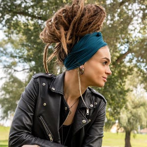 May include: A black leather jacket with silver zippers and buttons. The jacket is worn over a black top. A teal-colored headband is worn with brown dreadlocks. The person is wearing silver hoop earrings and a necklace.