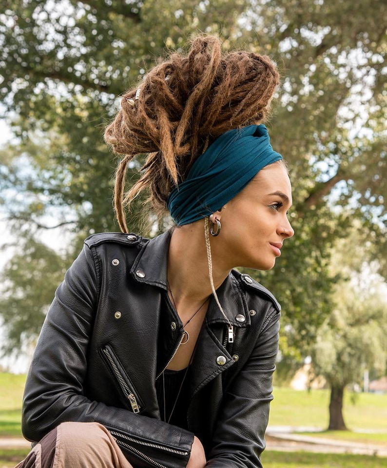 May include: A black leather jacket with silver zippers and buttons. The jacket is worn over a black top. A teal-colored headband is worn with brown dreadlocks. The person is wearing silver hoop earrings and a necklace.