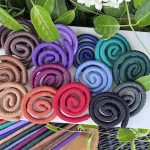 Puede incluir: Una colecci&oacute;n de adornos en forma de espiral hechos de cord&oacute;n cubierto de tela, presentados en una variedad de colores que incluyen marr&oacute;n, morado, azul, verde azulado, rojo y negro, dispuestos sobre una superficie blanca con hojas.