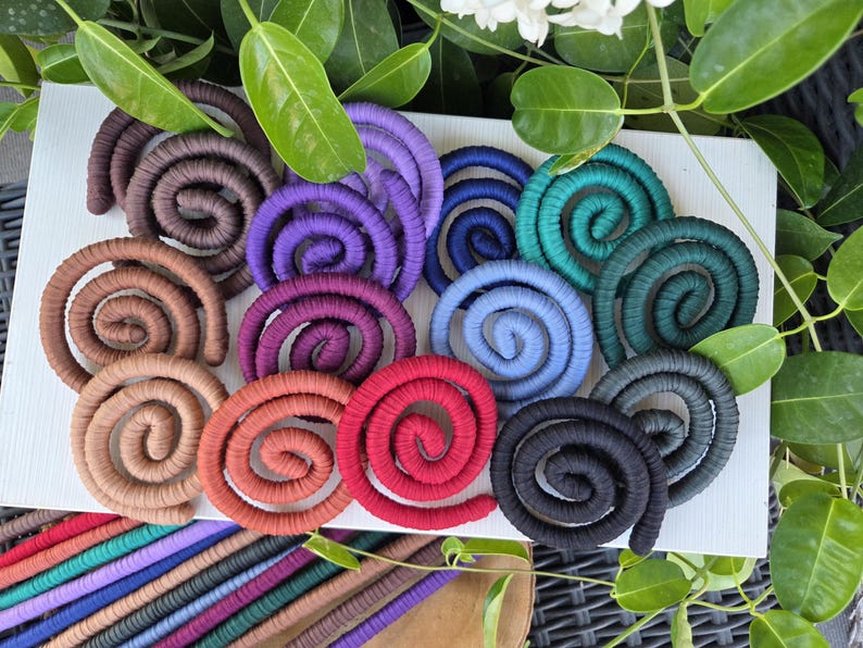 Puede incluir: Una colecci&oacute;n de adornos en forma de espiral hechos de cord&oacute;n cubierto de tela, presentados en una variedad de colores que incluyen marr&oacute;n, morado, azul, verde azulado, rojo y negro, dispuestos sobre una superficie blanca con hojas.