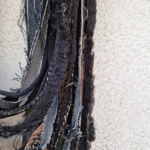 Pode incluir: Um close-up de um cachecol de tecido preto, marrom e dourado com bordas desfiadas. O cachecol &eacute; feito de v&aacute;rias tiras de tecido tecidas juntas.