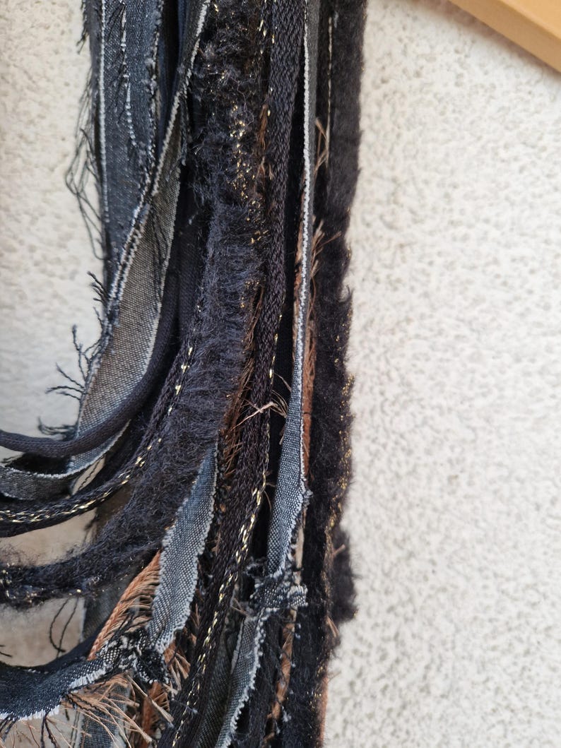 Pode incluir: Um close-up de um cachecol de tecido preto, marrom e dourado com bordas desfiadas. O cachecol &eacute; feito de v&aacute;rias tiras de tecido tecidas juntas.
