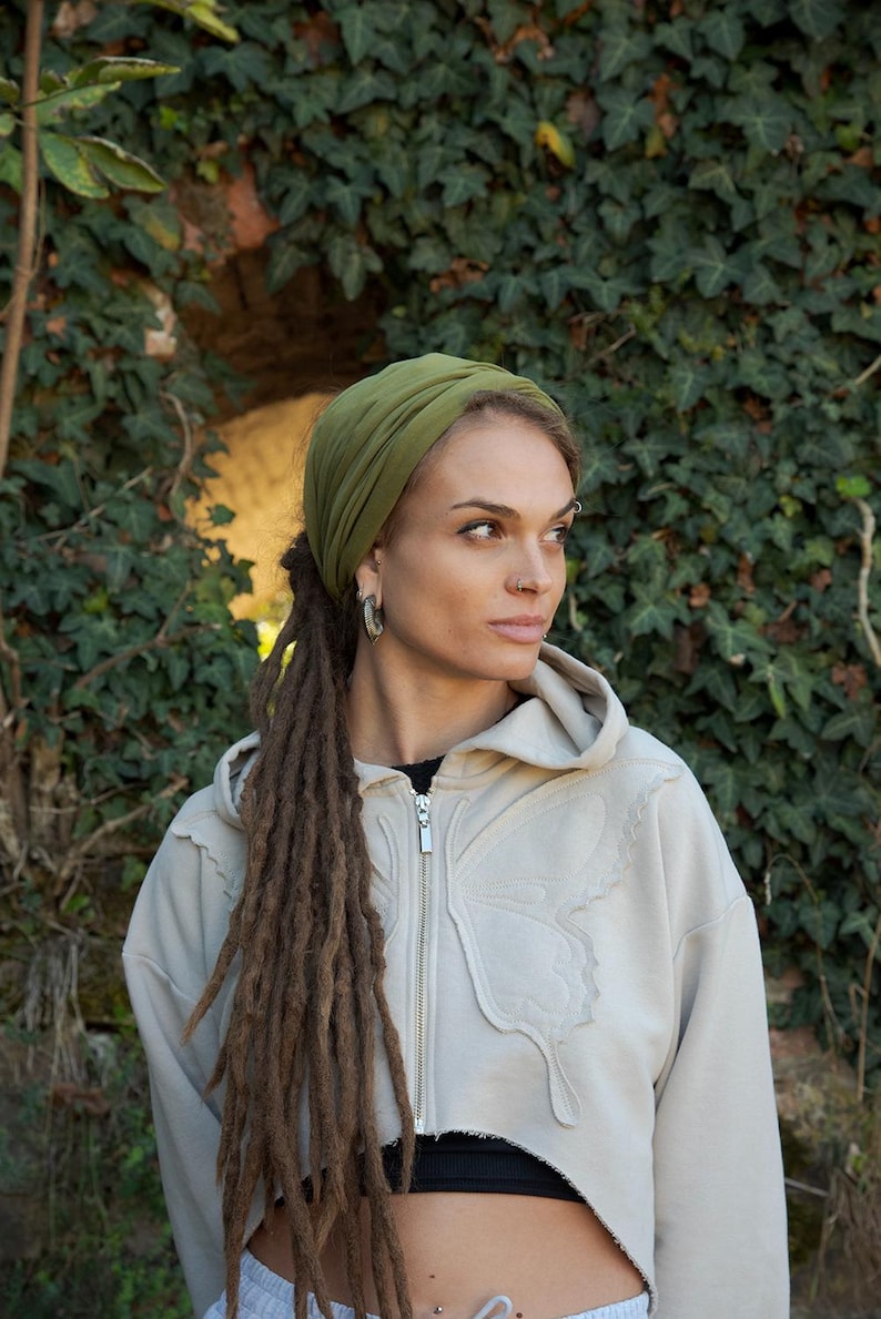 May include: A cropped beige zip-up hoodie featuring a stitched butterfly wing design on the front, paired with an olive green head wrap. The hoodie has a silver-toned zipper and a raw-edge hem, worn over a black top and light-colored bottoms.