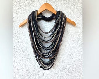 May include: A multi-strand statement necklace featuring layers of black, grey, and brown fabric strips and textured yarn. The strands are gathered at the top with a black wrapped cord, creating a cascading V-shape design that hangs from a wooden hanger.