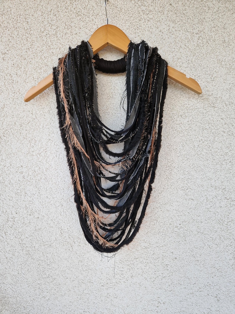 Pode incluir: Um cachecol longo e em camadas feito de tecido preto e marrom com bordas desfiadas. O cachecol est&aacute; pendurado em um cabide de madeira.