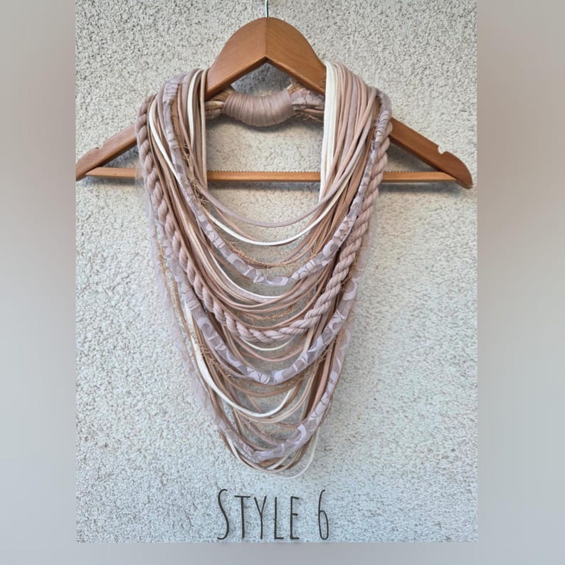 Op de afbeelding: Een beige infinity sjaal met meerdere texturen, waaronder gevlochten en gladde stoffen, en delicate kanten accenten. De sjaal wordt getoond op een houten hanger tegen een getextureerde muur. De tekst "STYLE 6" staat onderaan.