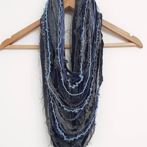May include: An infinity scarf constructed from multiple layered strips of dark blue denim, charcoal grey fabric, and light blue yarn. The design features frayed, raw edges throughout, creating a textured, multi-strand appearance hanging in loops.