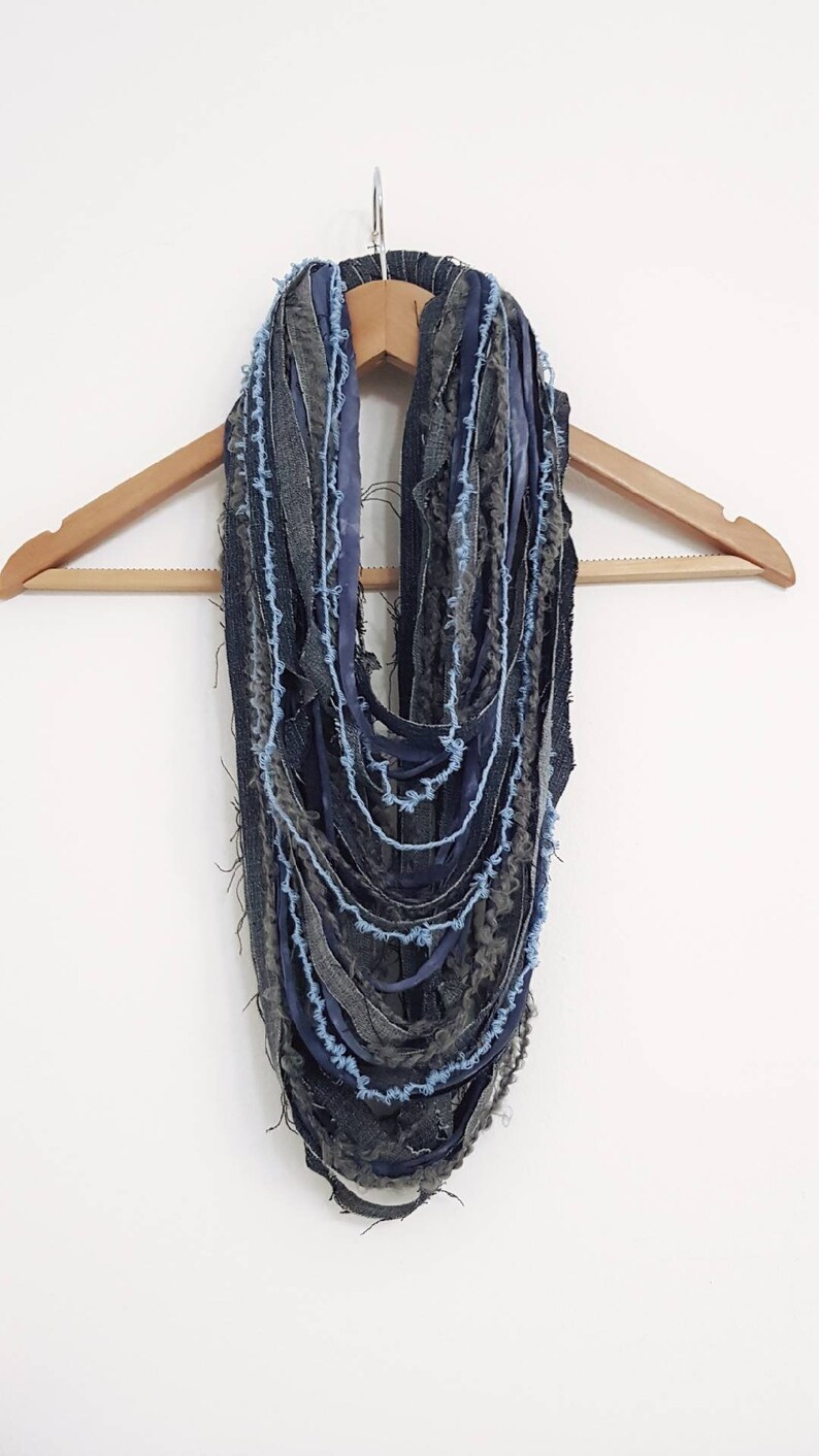 May include: An infinity scarf constructed from multiple layered strips of dark blue denim, charcoal grey fabric, and light blue yarn. The design features frayed, raw edges throughout, creating a textured, multi-strand appearance hanging in loops.