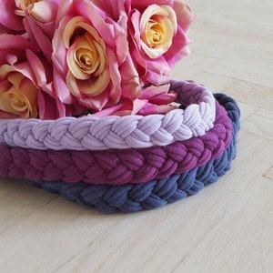 May include: Three braided headbands stacked together in light lavender, deep magenta, and slate blue colors. The headbands feature a thick, woven fabric texture. A bouquet of pink and yellow artificial roses rests behind the headbands.