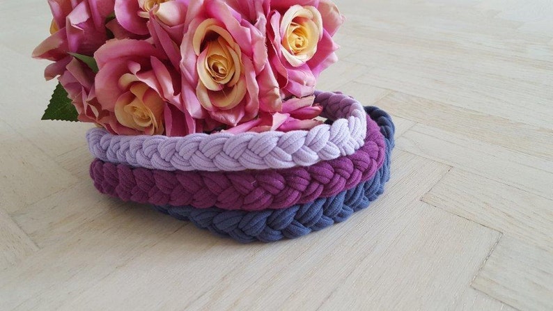May include: Three braided headbands stacked together in light lavender, deep magenta, and slate blue colors. The headbands feature a thick, woven fabric texture. A bouquet of pink and yellow artificial roses rests behind the headbands.