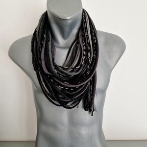 May include: A multi-strand infinity scarf featuring a layered design of fabric strips in shades of black, charcoal gray, and light gray. The scarf consists of numerous thin, rolled fabric loops that create a textured, voluminous appearance.