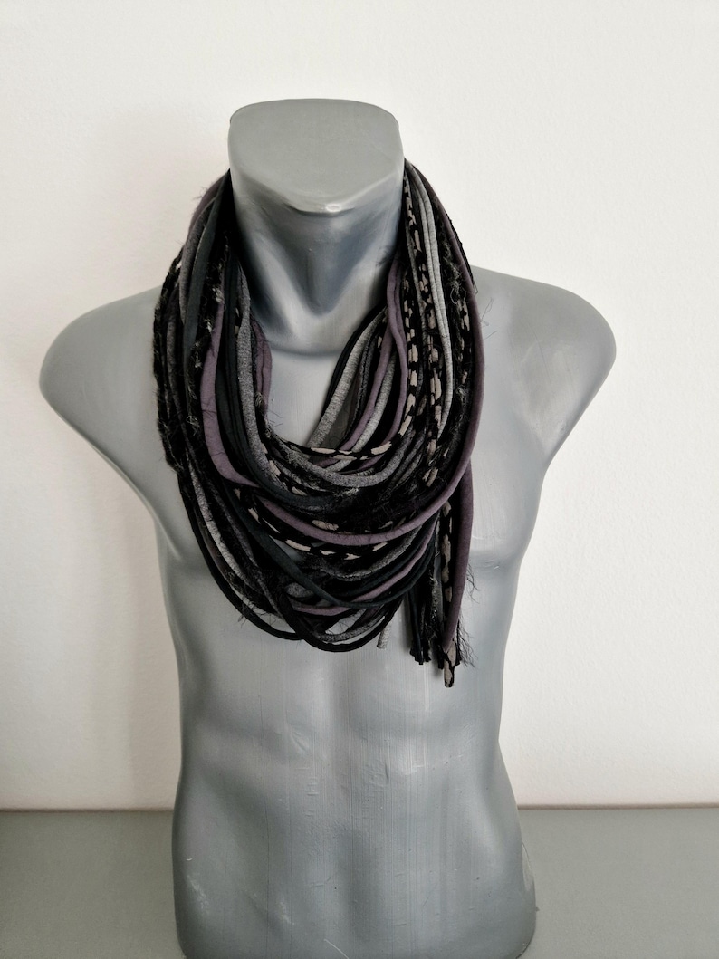 May include: A multi-strand infinity scarf featuring a layered design of fabric strips in shades of black, charcoal gray, and light gray. The scarf consists of numerous thin, rolled fabric loops that create a textured, voluminous appearance.