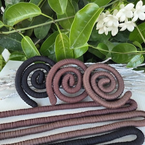 Puede incluir: Una colecci&oacute;n de formas en espiral flexibles cubiertas de tela en negro y varios tonos de marr&oacute;n. Estos objetos largos y cil&iacute;ndricos est&aacute;n enrollados en formas circulares en la parte superior, con longitudes rectas y paralelas que se extienden hacia abajo.